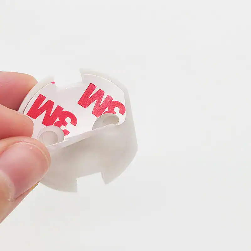 אדם מחזיק חפץ פלסטיק עגול עם גב דביק. לדבק יש לוגו אדום "3M" הנראה לעין. לאובייקט יש שני חורים וחריצים מסביב לקצוות, על רקע לבן רגיל.