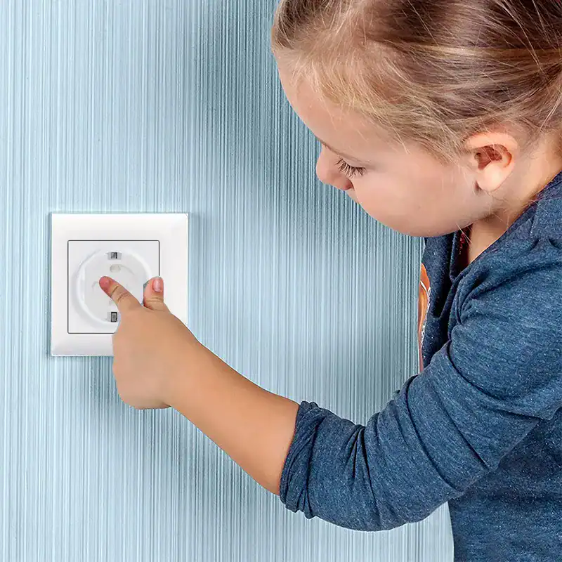 ילד צעיר מושיט יד לעבר שקע חשמל על קיר תכלת. הילד לובש חולצה כחולה כהה עם שרוולים ארוכים.